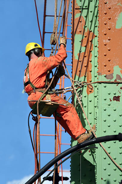 "Bridge worker lowers himself into position to perform maintenance high up on a suspension bridge. Good detail of safety equipment in use, literally providing job security."